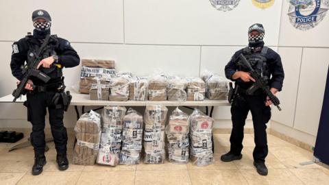 Two armed police officers standing either side of a collection of large packages of cocaine