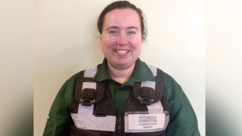 A woman with tied back dark hair is dressed in a green shirt and a black vest with high viz panels.