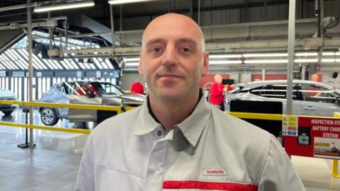 Jamie Whillians. He is standing in front of a production line of cars in the Nissan factory at Sunderland. His upper half is visible, and he is wearing a grey button up, with a Nissan logo on his left-hand side.