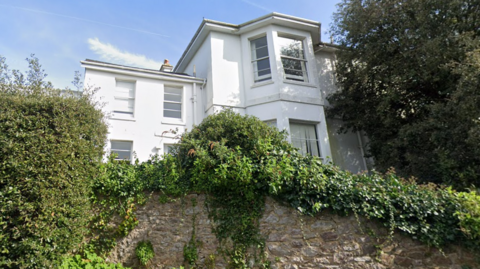 A shot of a big white house looking up from the road below. There is a big wall blocking the downstairs of the property with lots of ivy and greenery on top of the wall. There is a large tree to the right of the images the same height as the building and clear blue skies. 