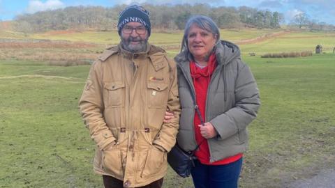 Man in black hat with glasses and beige coat stands arm in arm with woman. She is wearing a grey coat, red jumper and has grey hair.