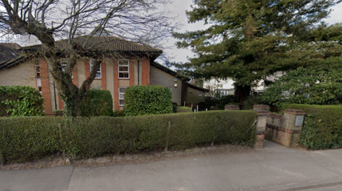 A general view of the two-storey GPs surgery, which is on a main road and behind a bush. A walled walkway is on the right of the surgery, leading from a pavement 