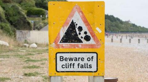 A sign on a shingle beach saying beware of cliff falls with gently sloping cliffs covered in greenery in the background