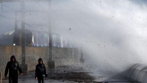 Two men, wearing black jackets with their hoods up being covered with water from a large white wave.