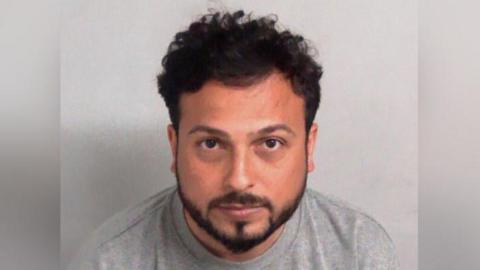 A man looks directly into the camera. He has curly dark hair and a neat beard. He wears a grey top and is not smiling