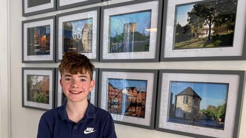 Isaac stood in front of a white wall with eight picture frames behind him with his photography in them. All the pictures have black frames in two rows of four. Isaac has short brown hair and is wearing a navy blue polo t-shirt.