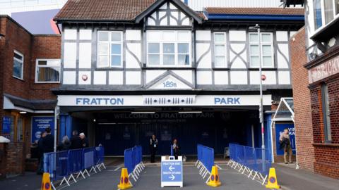 A generic photo taken previously shows blue barriers lined up outside the entrance to Fratton Park, with Fratton Park written in blue above on the front of the building