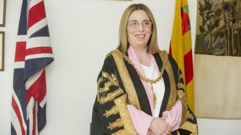 Jeanette Whalen smiling to the right of the camera. She has shoulder length blonde hair and is wearing thin-framed glasses. She is wearing a black-and-gold ceremonial robe and a gold chain around her chest. She is wearing a baby pink blazer and white top underneath. There is a Union Flag behind her and the yellow-and-red Carlisle flag.