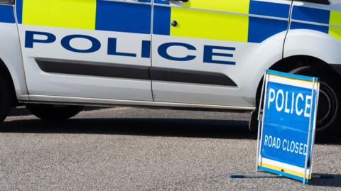 Side of a police car with a POLICE Road Closed sign in front of it.