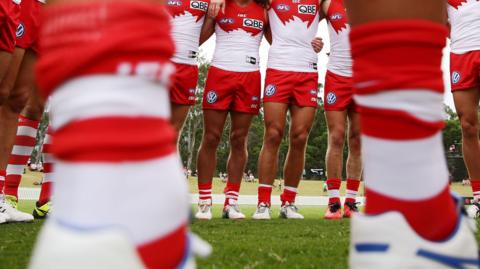 A pair of blurry legs in the foreground with a line of Sydney Swans players in the background
