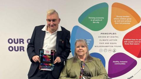 A man with blond hair stands next to a woman in a wheelchair next to him. He holds a black piece of paper with multi-coloured writing on it, saying 'The best of the best' and 'Finalist' on it.