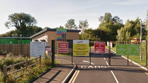 The closed gates of Bourne Household Waste Recycling Centre which are covered with various signs. In the background is a small building with a slanted black roof. On the left is a wooden fence and some grassland