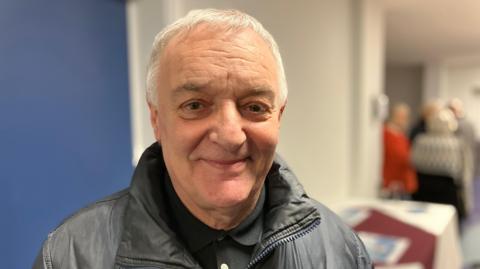 Lou Macari, a man with grey hair, is wearing a black leather jacket and a black shirt. He is standing in a well-lit room with a group of people in the background.