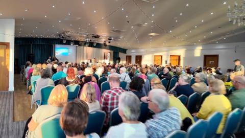 A large group of people say in a room while a meeting takes place. Guests are sat on blue chairs and a projection screen has been set up at the front of the room with four people on the stage.