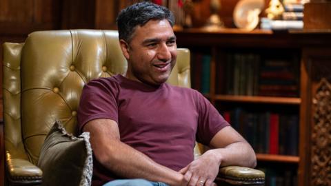 Nick Mohammed is sitting in a large leather armchair. He is a youngish man with dark hair and a bit of stubble and wearing a dark red top with jeans.