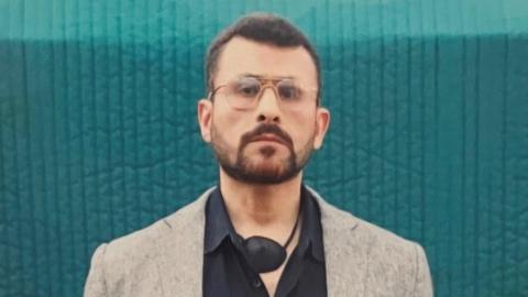 A head-to-chest shot of Abu Zubaydah, a man wearing glasses and with a defined beard, carrying an eye-patch around his neck. He is wearing a black shirt and grey blazer.