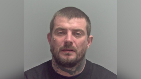 A mugshot image of Ryan Leggett. He is wearing a black T-shirt and has tattoos on his neck. He has a piercing under his right eye. He is looking into the camera.