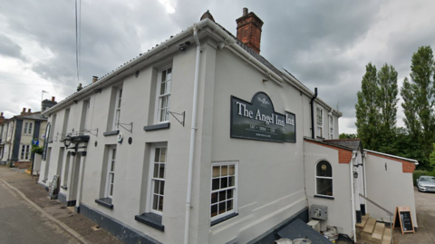 A general view of the exterior of The Angel Inn in Wangford, Suffolk. It shows a grey building with the sign detailing the name of the pub on the sign. Beer kegs can be seen lying outside the entrance to the pub cellar.