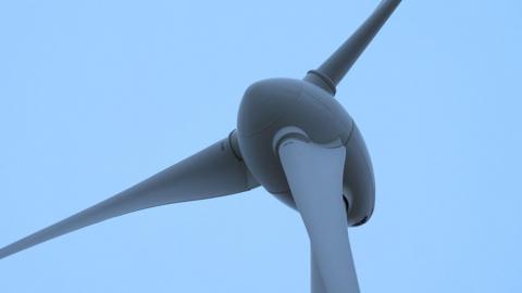 A close up of the centre of a wind turbine. Three white blades are attached to the dome shaped centre which stands on top of a large mast.