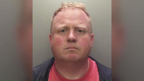Robert Bush with short, light brown hair is shown in a straight‑on mugshot photograph. He has a neutral expression and is wearing a dark jacket over a red shirt. The background is plain and grey.