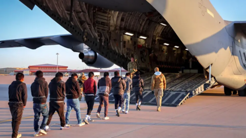 A queue of men with chains around their waists walk into a US military plane