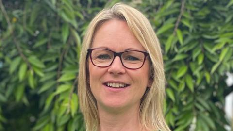 A head shot of Hazel Boyce. She has blonde hair, is wearing glasses and there are green leaves in the background of the photo. 