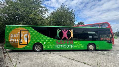 A view of the side of the bus. The bus is green and has stickers promoting Safer Plumouth