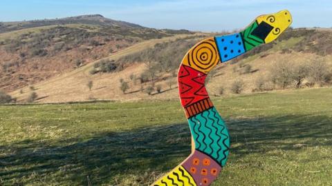 A large wooden snake painted in various bright colours and patterns. It is nailed to a wooden stick with an information sign and a QR code beneath it. In the background you can see the Mendip Hills on a sunny day.