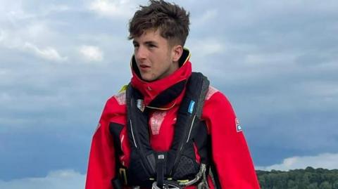 Image of Thomas Roy he is wearing red waterproofs and a black life vest at the helm of a boat