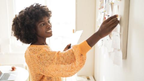 Smiling woman sticking note on board while holding digital tablet at home office - stock photo