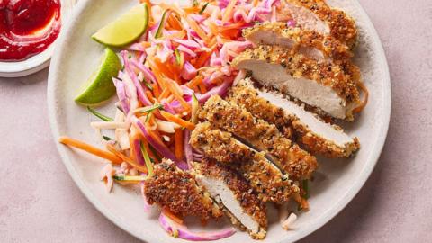 A breadded chicken breast sliced up on a plate with colourful slaw on a pale pink surface