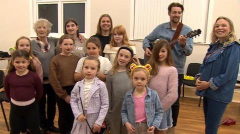 A group of 10 girls aged between seven to 13 years stand in four rows with four adults in the back row. They are in the village hall facing the camera. One child wears Pudsey Bear ears. One of the adults has a guitar.