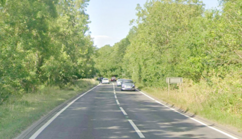 Google Maps images of the A509 near Little Harrowden. It shows a grey road with white dashed markings in the middle, and two solid lines at either side. The road is bordered by tall green foliage and a light blue sky can be seen at the top of the image. There are several cars on the road in both directions in the distance.