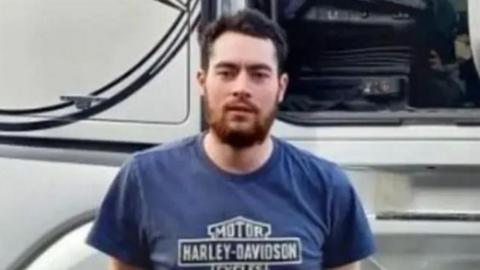 Gregory Newcombe wearing a blue t-shirt that says "Harley-Davison" on it stands in front of a white lorry