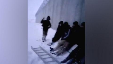 A number of migrants are seen sheltering against the wall in the snow, with a ladder at their feet - in a still from a video supplied to the BBC by a people smuggler