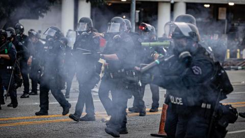 Members of Los Angeles Police Department in riot gear walk down the street, smoke around them