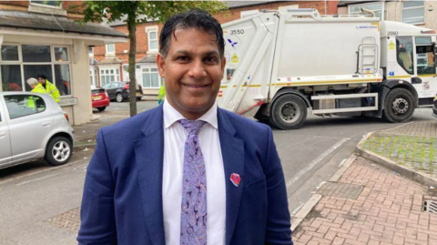 Councillor Majid Mahmood wearing a blue suit jacket and urle tie stands outdoors and looks at the camera.