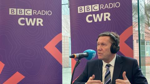 A man wearing a suit sits at desk, speaking into a microphone. Purple boards with the BBC Radio CWR logo and text can be seen behind him.