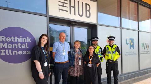 Three women and one man stand outside a building alongside two male police officers. The officers are in black outfits with black police hats and yellow fluorescent police vests. There is a sign above the door that reads "the hub".