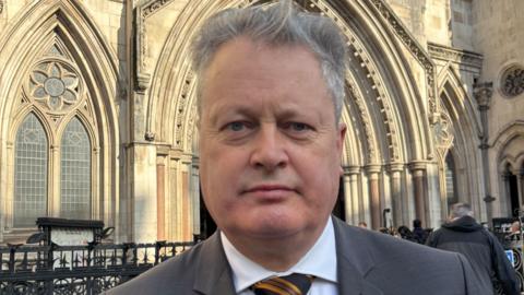 Vincent Kearney, a blue-eyed, grey-haired man in a grey suit, white shirt and orange and black striped tie. He is in front of an ornate stone building.