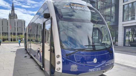 A blue and white rapid transit vehicle branded “Aberdeen Rapid Transit” is parked in the city-centre under a bright, partly cloudy sky. It is in front of the City's Town House. 