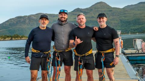 Four men in harnesses and athletic clothing stand together on a dock beside calm water, with green hills and mountains behind them.