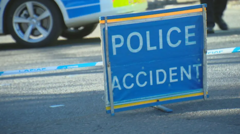 A blue police accident sign in front of police tape and the wheel of a police car.