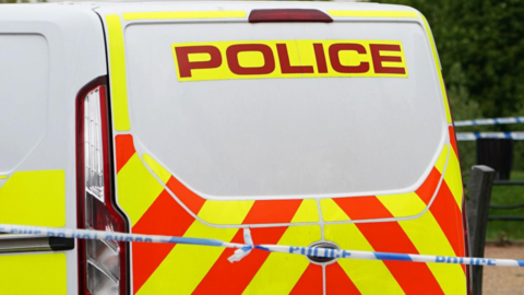 A white police van with red and yellow marking and a blue and white police tape in front