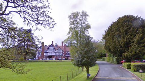 A driveway leading up to a red brick building with tudor panelling. There is a large patch of grass in front of the building and to the left of the driveway. There are also several cars parked in front of the building. 