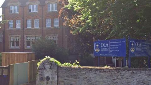 Outside Our Lady's Abingdon on  a sunny day. Blue school signs are installed in front of the building.
