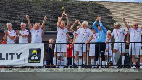 Member of the England men's over-75s football team standing on a balcony ion wehite footbal strip, lifting the world cup trophy