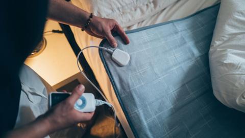  a hand holding the remote of an electric blanket, next to a bed with a blue electric blanket