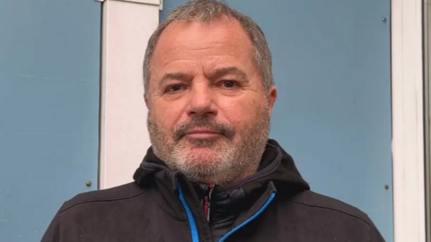 Andy Gavey has short dark hair and a short beard. He is wearing a black jacket