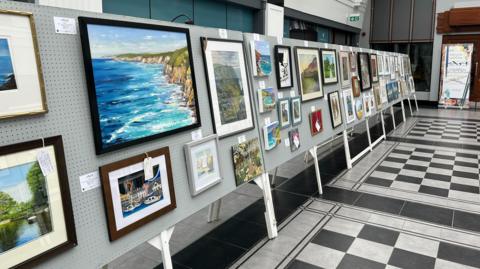 Artwork lines metal panels put up in a grand room with white and black tiles, one piece shows the rugged Manx coastline in a painting, another is a print of Peel harbour.